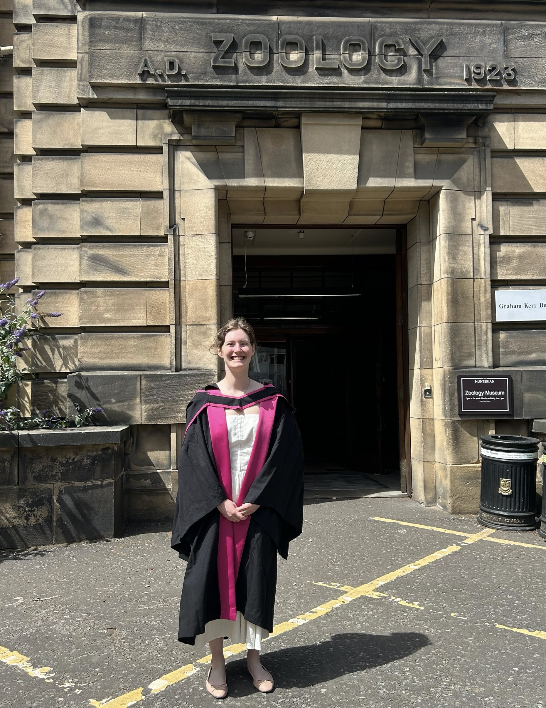 Rowan wearing the Glasgow University doctoral robe over a white dress, standing in front of the Graham Kerr zoology building.